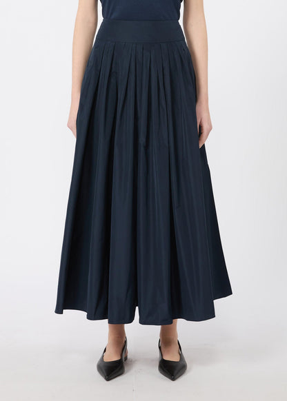 A model wearing a dark navy blue pleated midi skirt with a wide waistband, paired with black pointed-toe flats, shown against a plain light gray studio background.