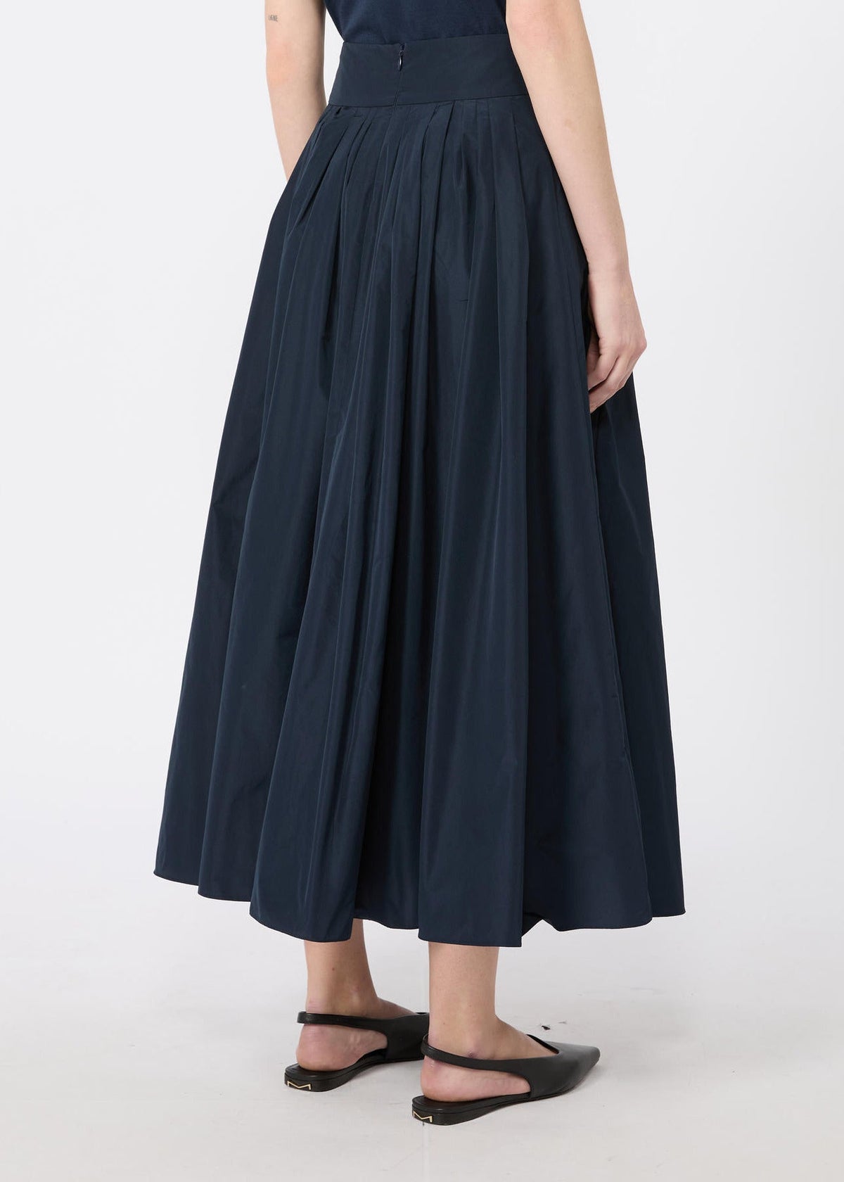A navy blue pleated midi skirt worn by a model, shown from the waist down against a light gray studio background.