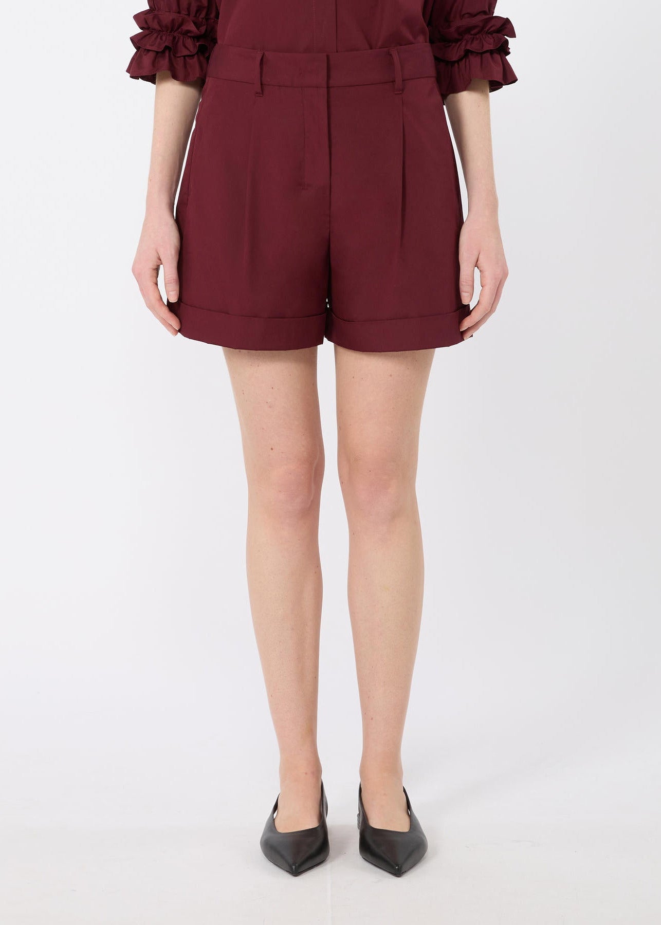 A model is shown from the waist down wearing high-waisted, pleated burgundy shorts with a matching ruffled top and black pointed-toe heels, set against a plain light gray studio background.