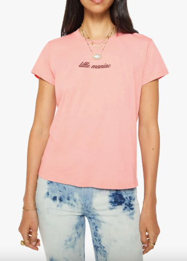 A woman wearing a light pink short-sleeve t-shirt with 'little maniac' printed in dark pink cursive on the chest, paired with light blue and white tie-dye jeans.