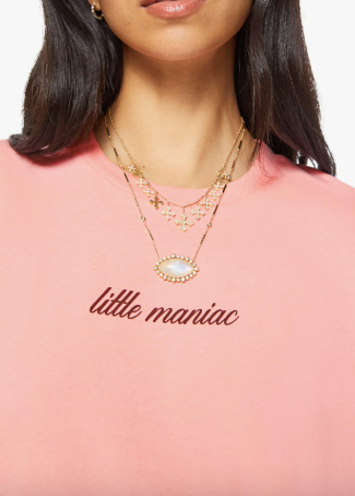 A close-up of a model wearing a light pink t-shirt with 'little maniac' embroidered in dark red across the chest, accessorized with layered gold necklaces.