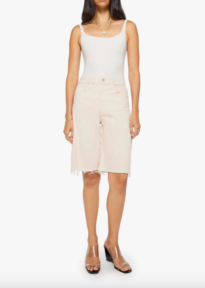 A model wearing a white ribbed tank top and light beige denim shorts with a raw hem, paired with clear-strap heeled sandals, against a plain white background.