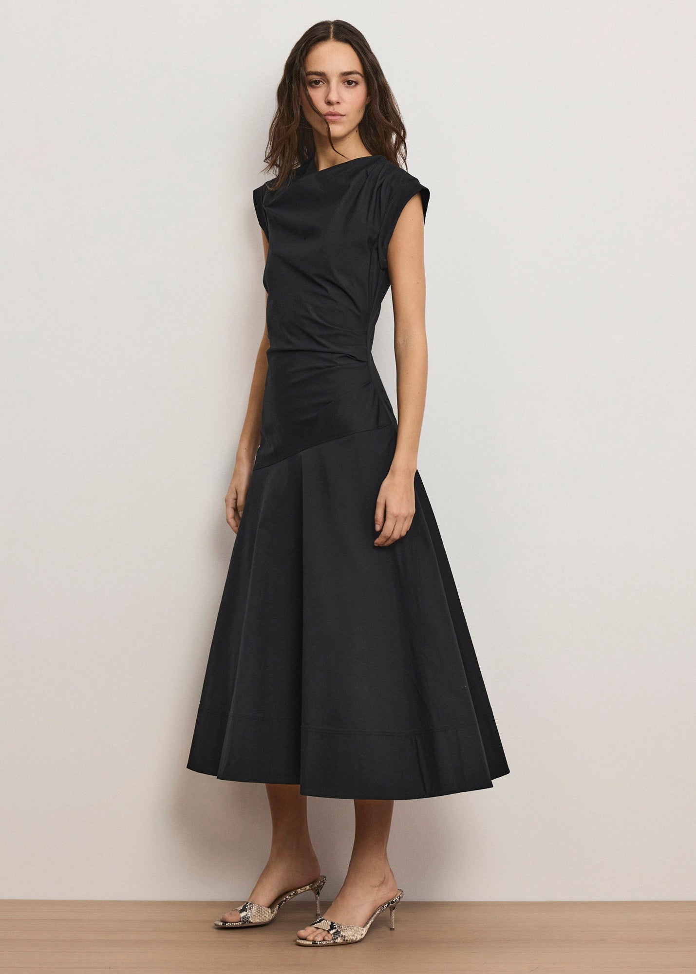 A woman modeling a sleeveless black midi dress with a gathered, draped bodice and a flared skirt, paired with metallic heeled sandals, set against a plain off-white wall.