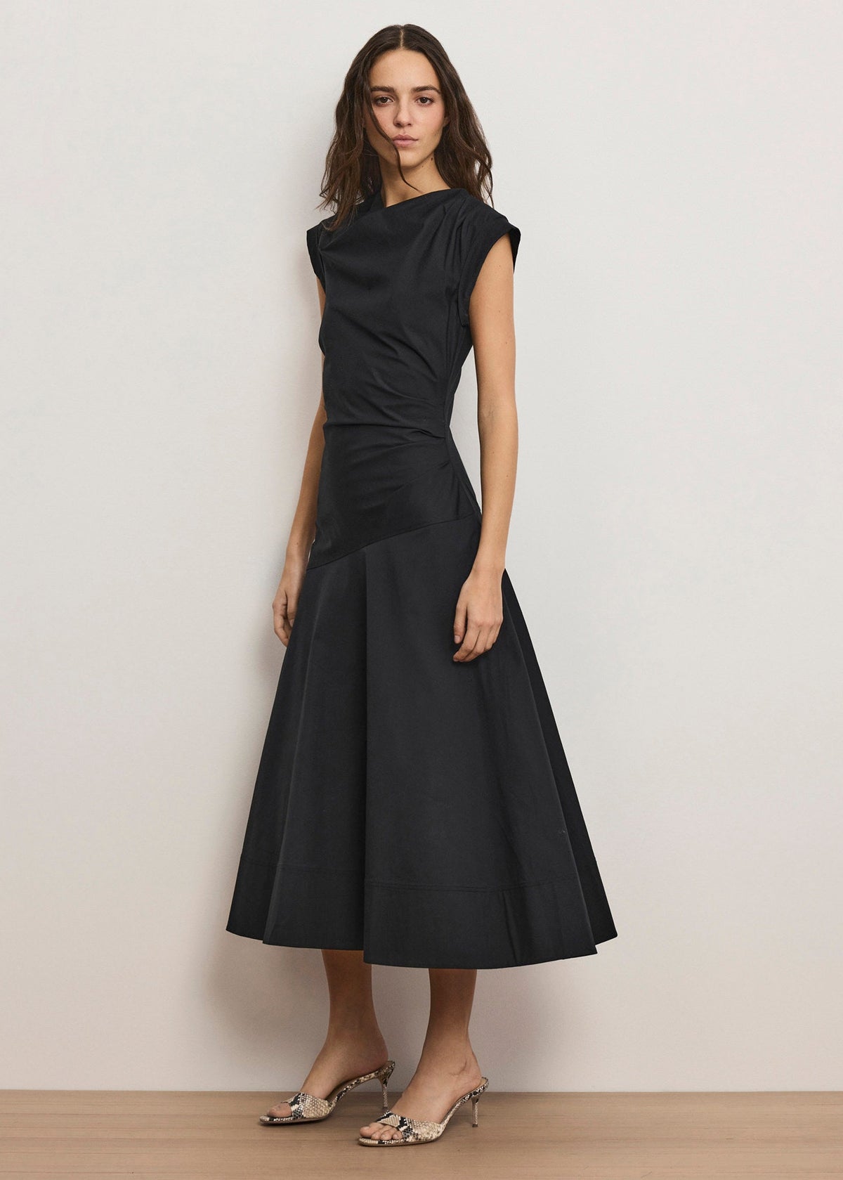 A woman modeling a sleeveless black midi dress with a gathered, draped bodice and a flared skirt, paired with metallic heeled sandals, set against a plain off-white wall.