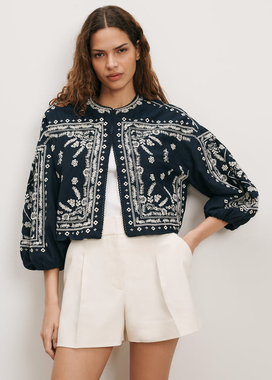 A woman modeling a cropped navy blue jacket with intricate white floral embroidery and silver beadwork, paired with cream pleated shorts against a light gray studio background.