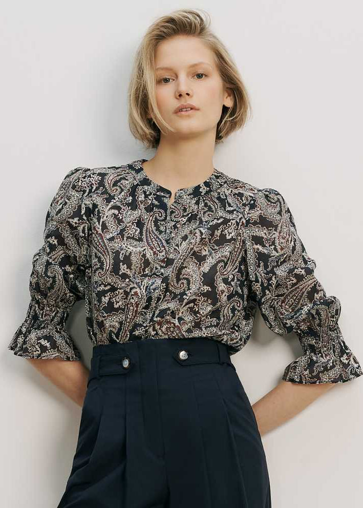 Woman wearing a patterned blouse and dark pants against a plain background