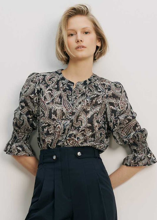 Woman wearing a patterned blouse and dark pants against a plain background