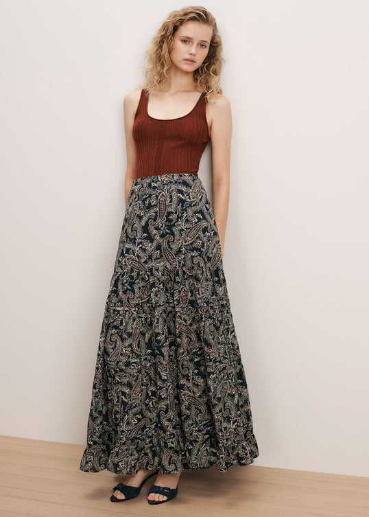 A woman stands against a plain off-white wall wearing a sleeveless rust-brown ribbed tank top and a long dark patterned maxi skirt with black sandals.
