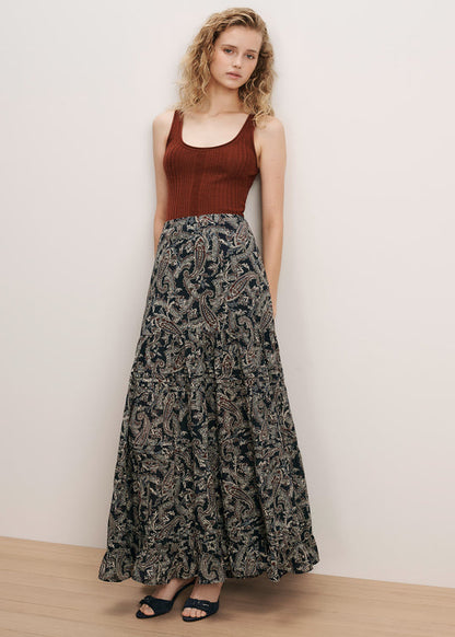 A woman stands against a plain off-white wall wearing a sleeveless rust-brown ribbed tank top and a long dark patterned maxi skirt with black sandals.