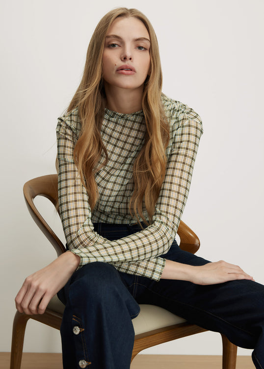 A woman with long light brown hair sits on a wooden chair wearing a beige and green plaid long-sleeve top and dark blue jeans against a plain off-white background.