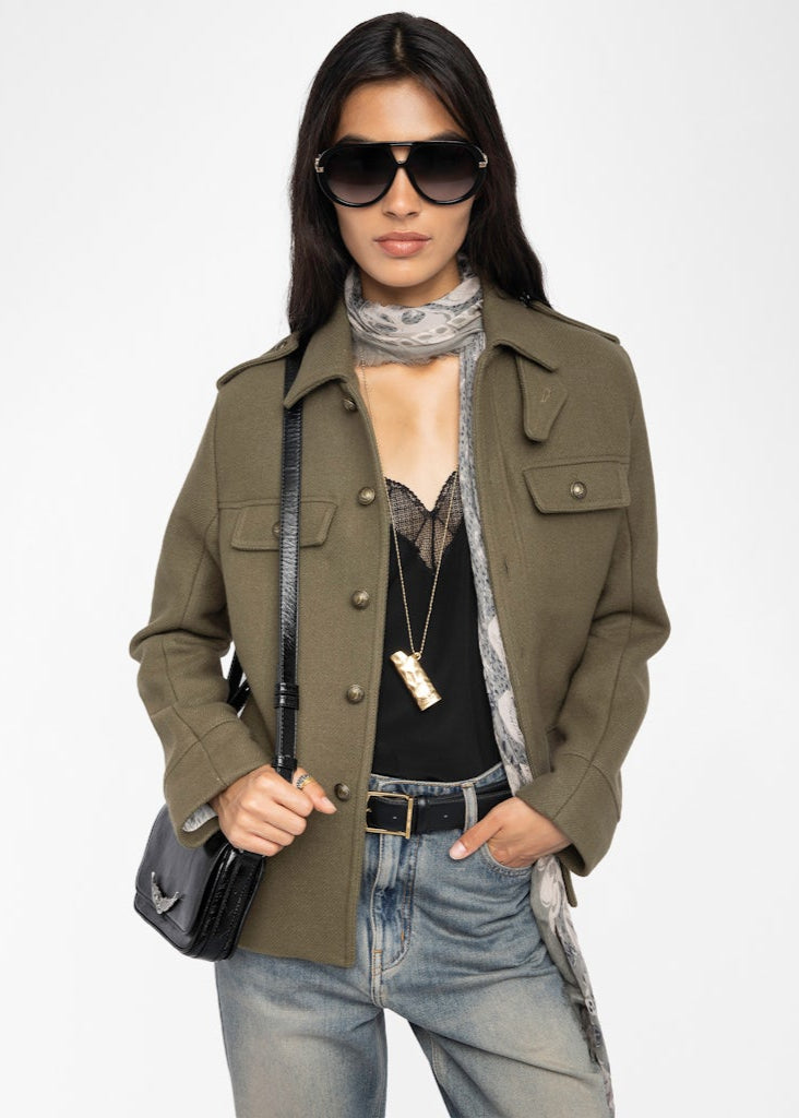 Woman wearing an olive green coat, sunglasses, and holding a black handbag on a white background
