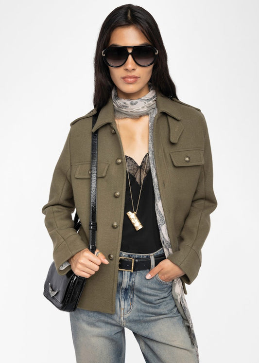 Woman wearing an olive green coat, sunglasses, and holding a black handbag on a white background