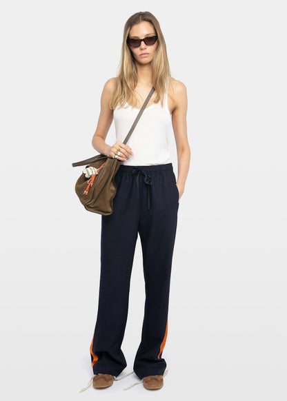 Woman wearing a white top and black pants with orange accents, holding a brown bag on a white background.