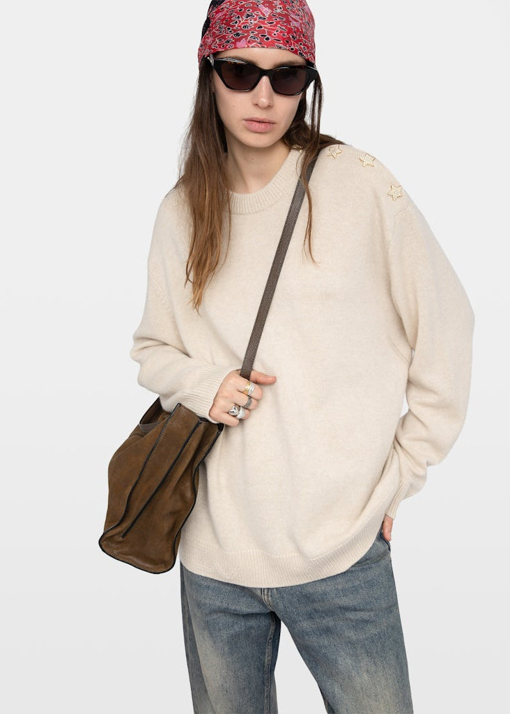 Person wearing a beige sweater, sunglasses, and a patterned headband, holding a brown bag on a white background.