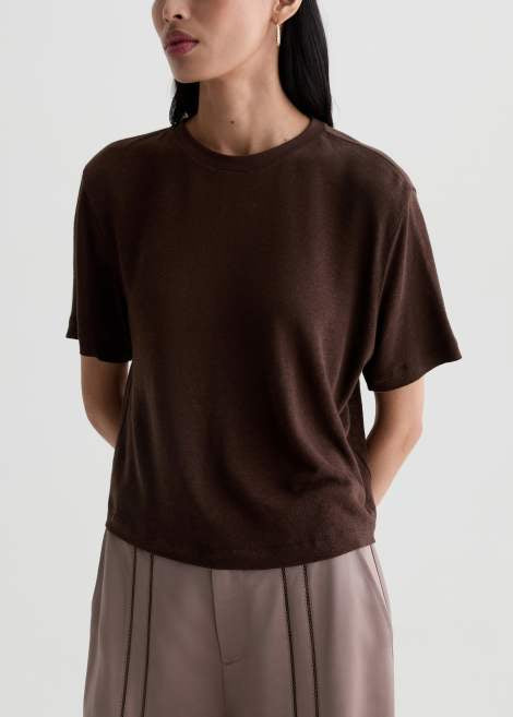 A woman wearing a dark brown short-sleeve crewneck knit top and light taupe pleated trousers, shown from the side against a light gray studio background.