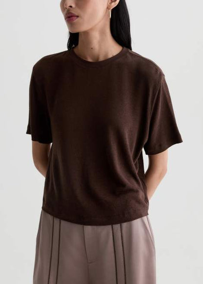 A woman wearing a dark brown short-sleeve crewneck knit top and light taupe pleated trousers, shown from the side against a light gray studio background.