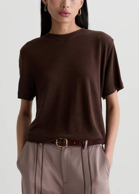 A woman wearing a dark brown short-sleeve crewneck knit top tucked into taupe pleated trousers with a brown belt, shown from the nose down against a light gray studio background.