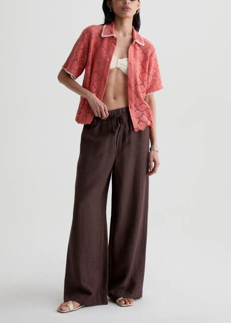 A model wearing a coral pink short-sleeve button-up shirt with a snakeskin pattern and dark brown wide-leg pants, styled with a white bikini top and flat sandals against a light gray studio background.