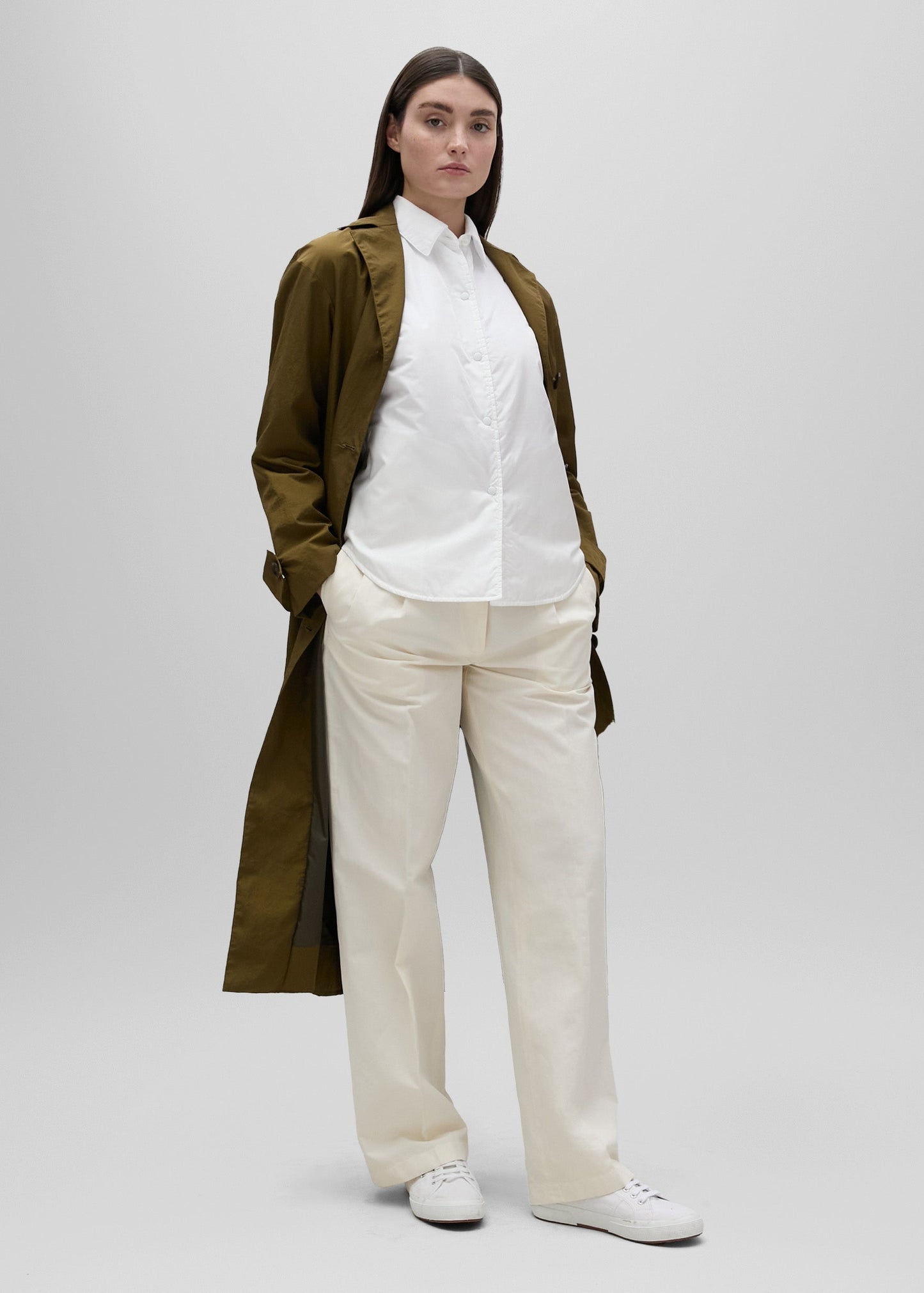 Person wearing a long olive green coat over a white shirt and beige pants on a light gray background