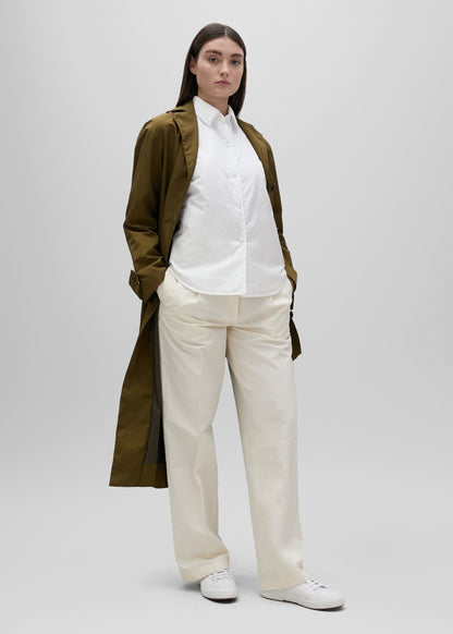 Person wearing a long olive green coat over a white shirt and beige pants on a light gray background