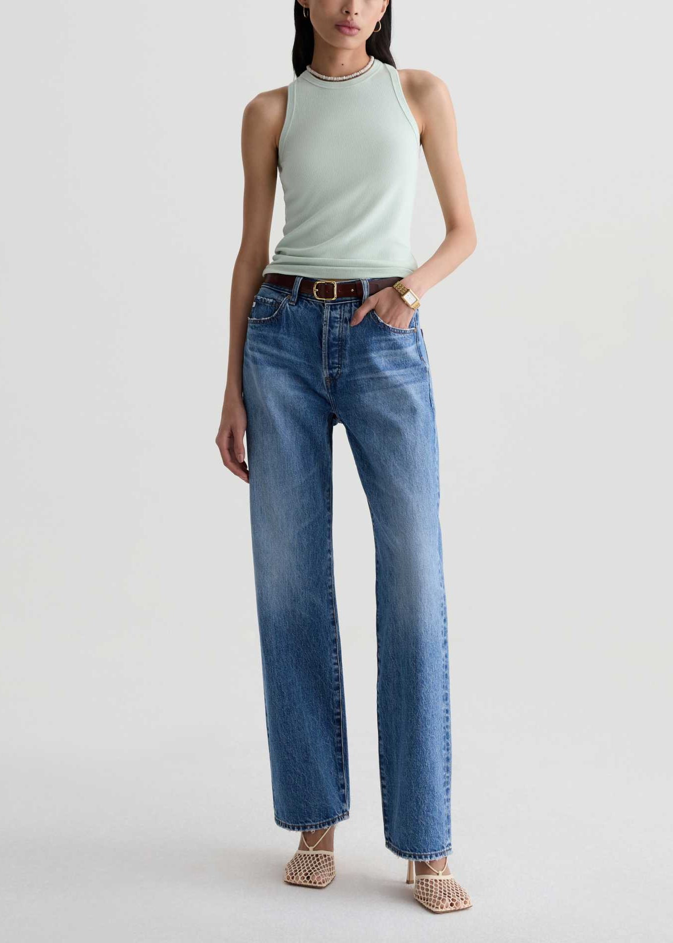 A model wearing a sleeveless pale green top and high-waisted, wide-leg blue jeans with a brown belt, standing against a light gray studio background.
