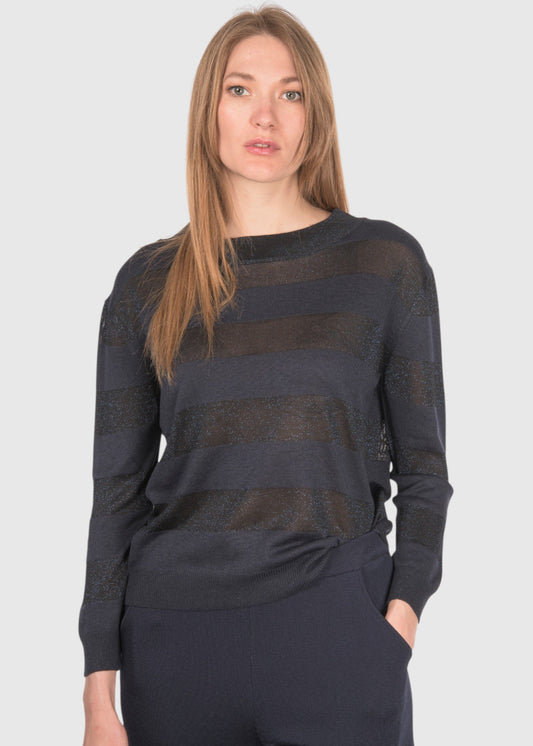 A woman modeling a dark charcoal gray and black horizontally striped knit sweater with a crew neck, paired with dark navy pants, against a solid light gray background.