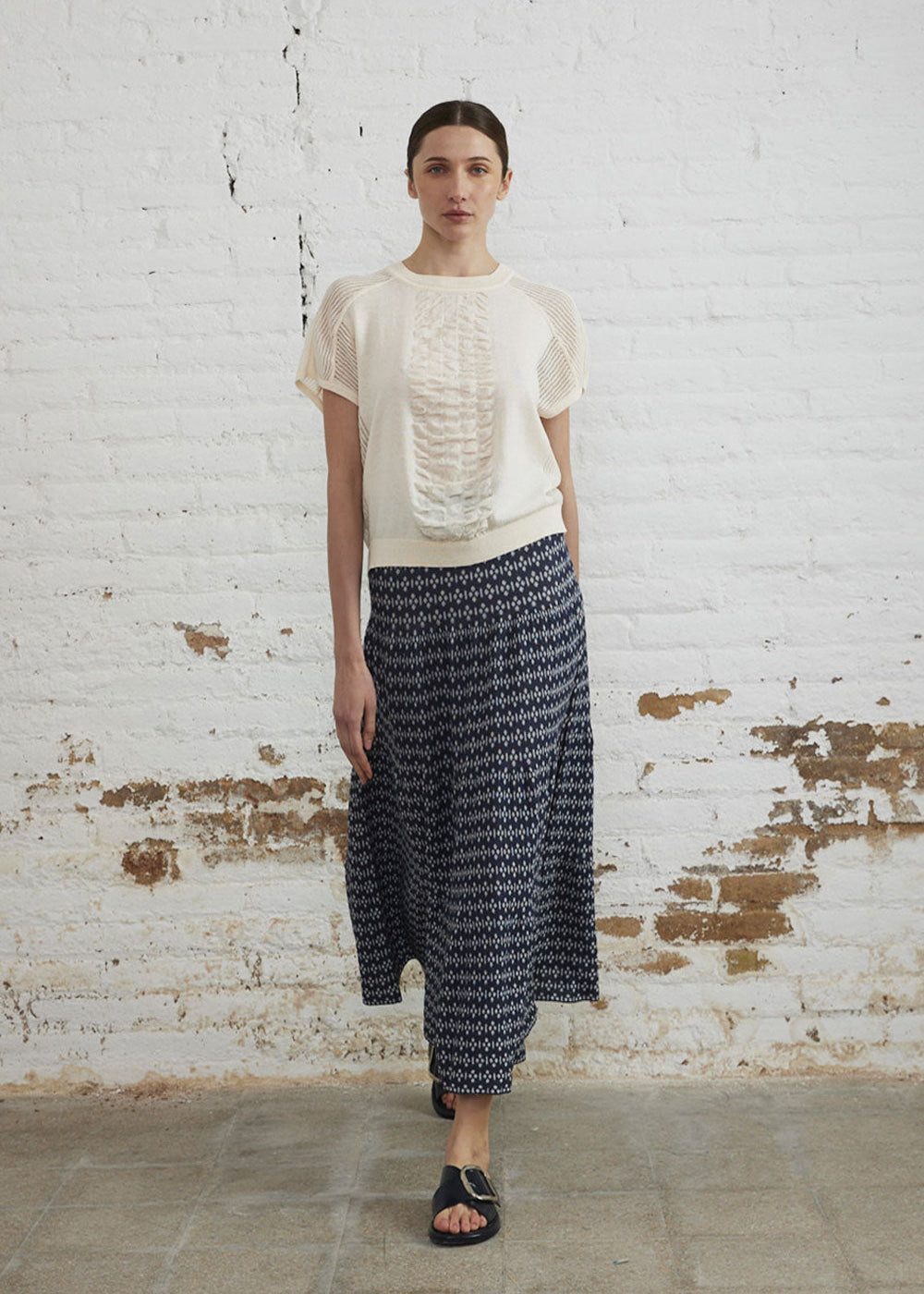 Woman wearing a white blouse and patterned skirt against a white brick wall.