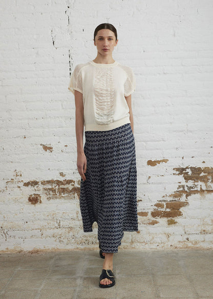 Woman wearing a white blouse and patterned skirt against a white brick wall.