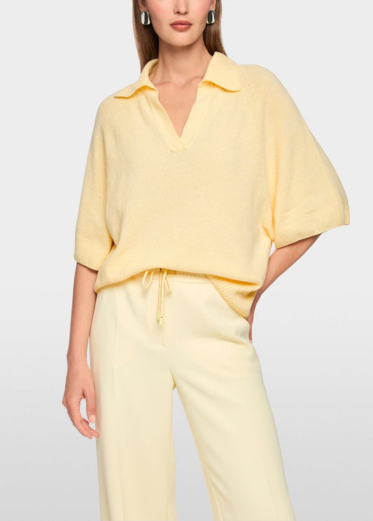 A woman wearing a pale yellow short-sleeve collared knit top and matching light yellow drawstring pants, photographed against a clean light gray studio background.