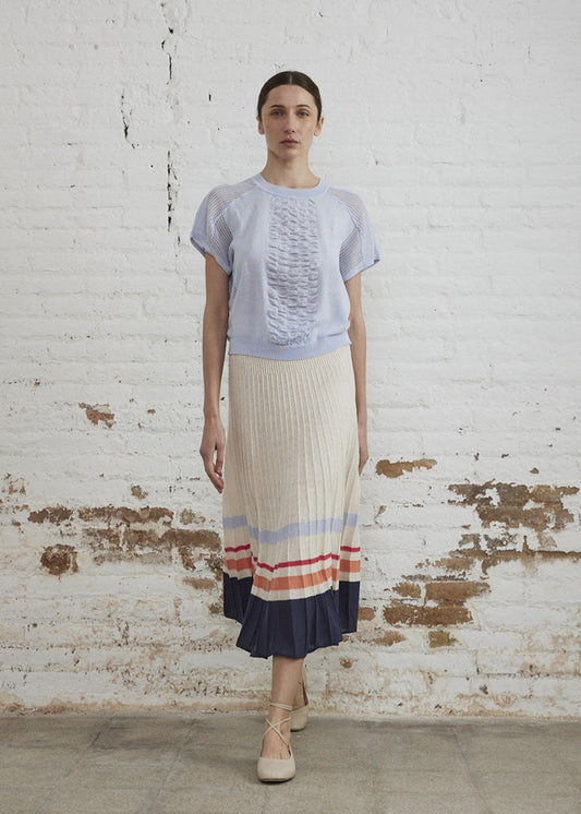 Woman wearing a light blue top and striped skirt against a white brick wall.